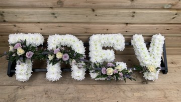 Trev - custom name funeral tribute based in whate crysanth with lilac and white sprays 