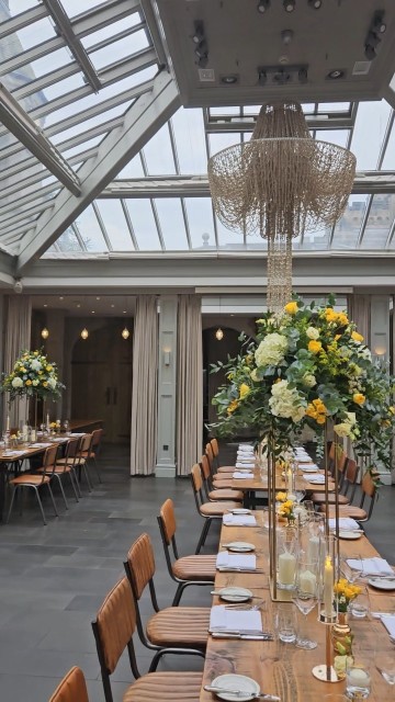 Banquette tablescape design at hampton manor for Mediterranean Shipping Company - Tall gold stands with large yellow and ivory flowers displays - short stone urns of flowers with gold and glass bud vases of flowers and gold and glass candle holders in between